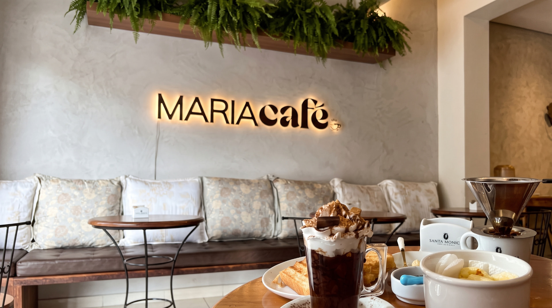 Salão do Maria Café com o letreiro iluminado, plantas, chocolate quente e café coado na mesa